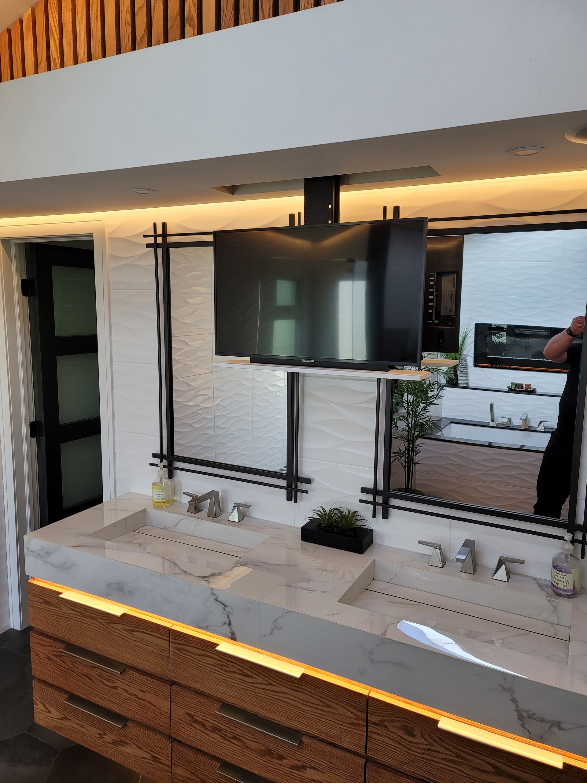 Modern bathroom vanity with dual sinks, large wall mirror, TV, and wood-accented lighting against white tile.