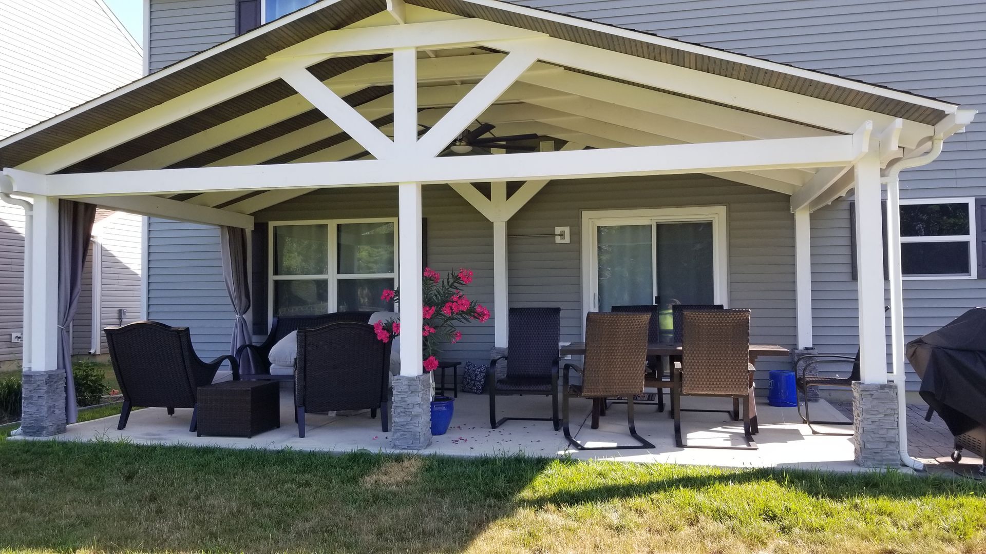 Covered patio with black chairs and brown table under a cream pergola in front of a house.