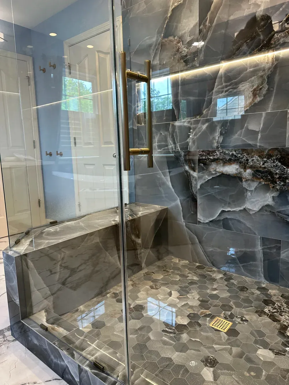 Modern glass shower with marble-look walls, hex tile floor, and brass handle in a bathroom