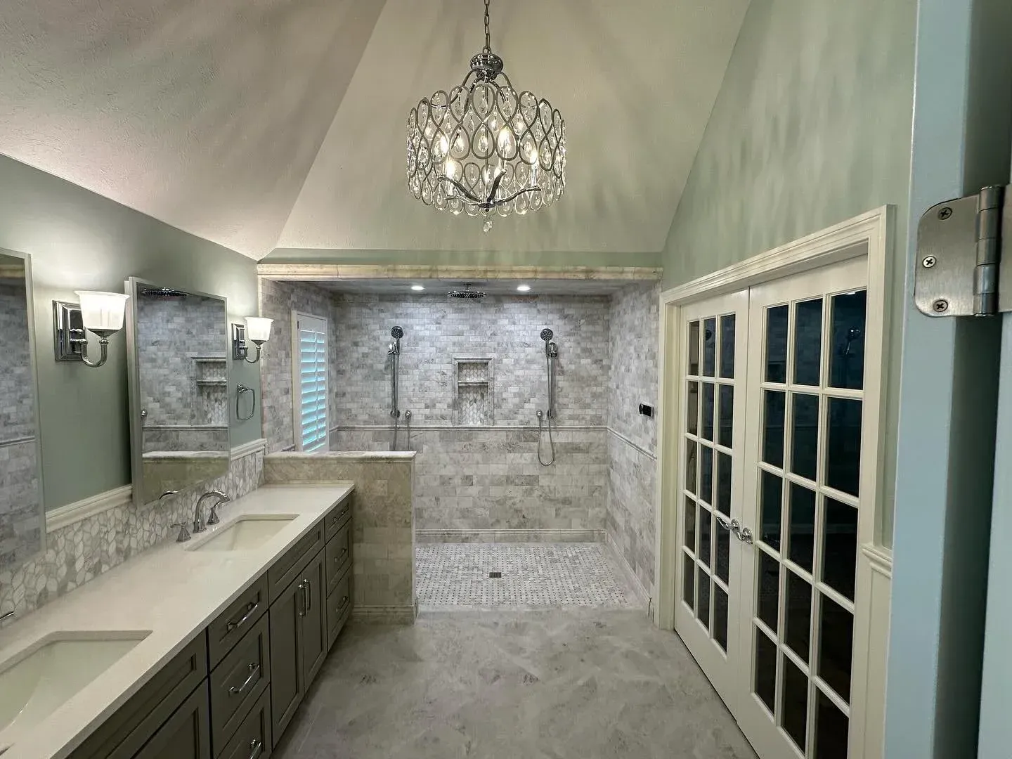 Elegant bathroom with chandelier, double vanity, and large walk-in shower in light stone and gray tones