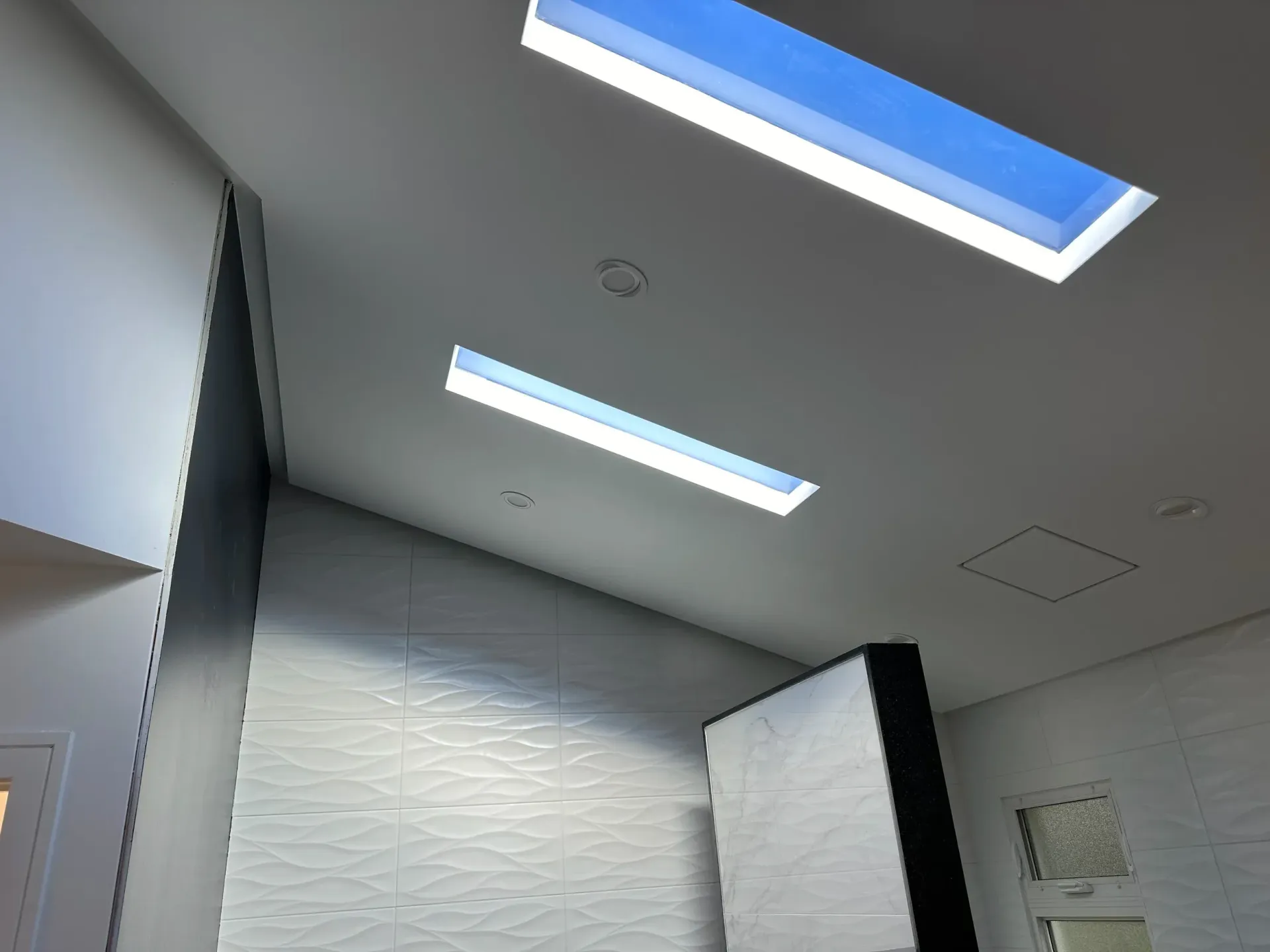 Modern bathroom with white tile walls, gray ceiling, and two skylights letting in blue daylight