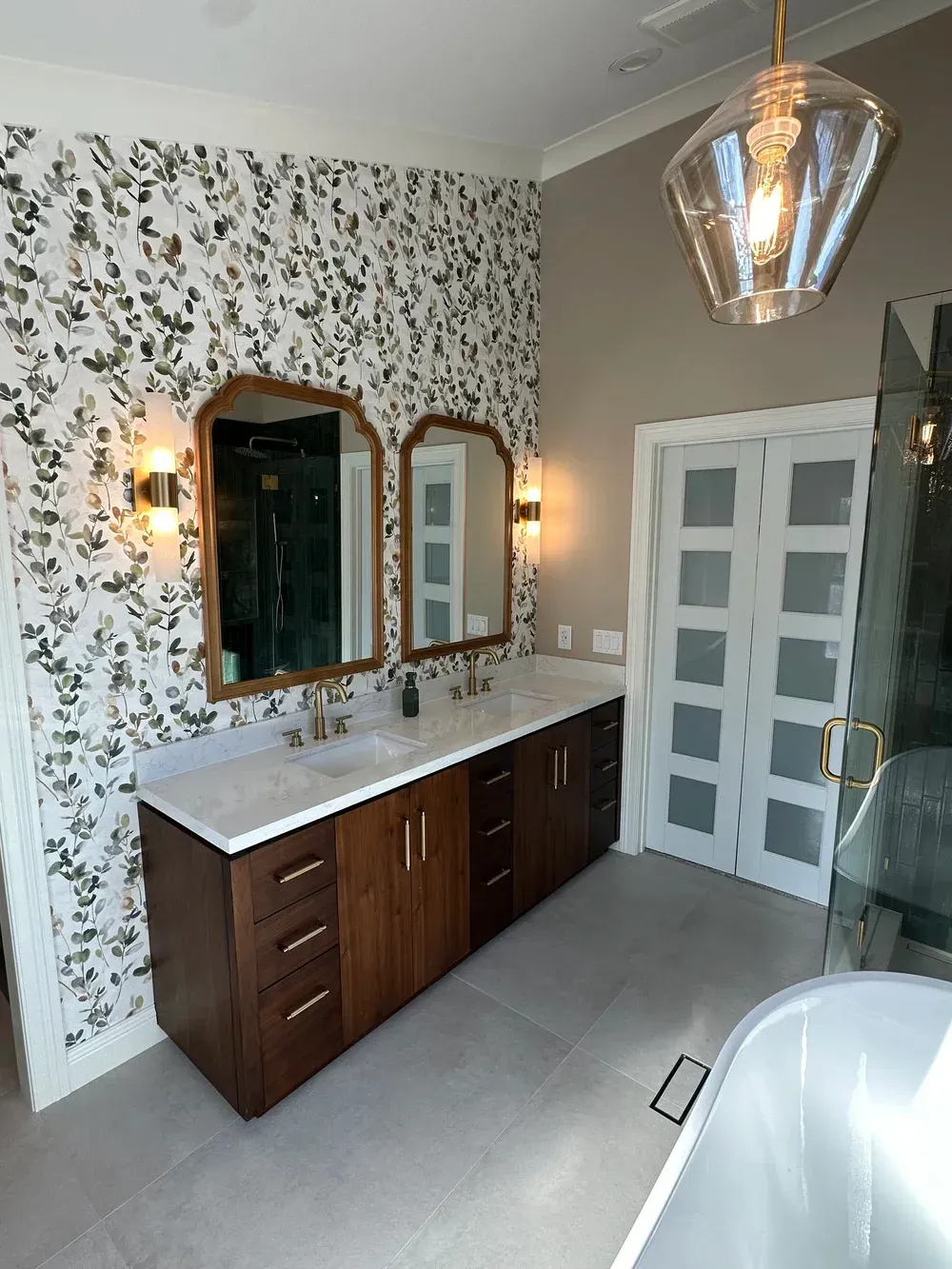 Vintage-style bathroom with floral wallpaper, double vanity, mirrors, pendant light, and white shower doors