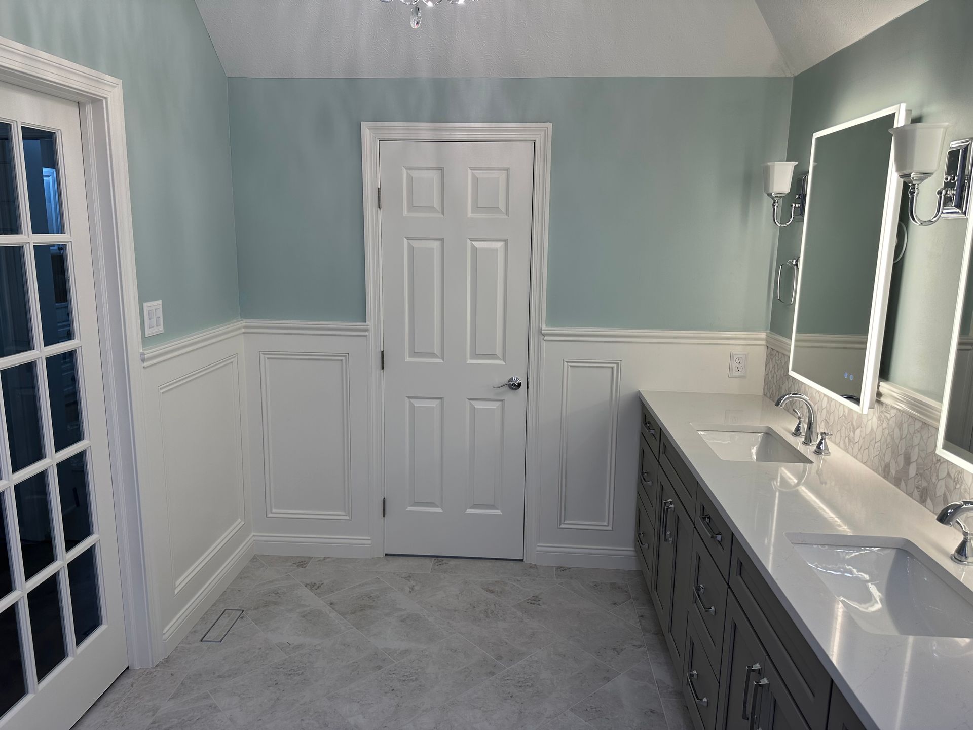 Bright bathroom with white wainscoting, blue-gray walls, double sinks, and a closed door.