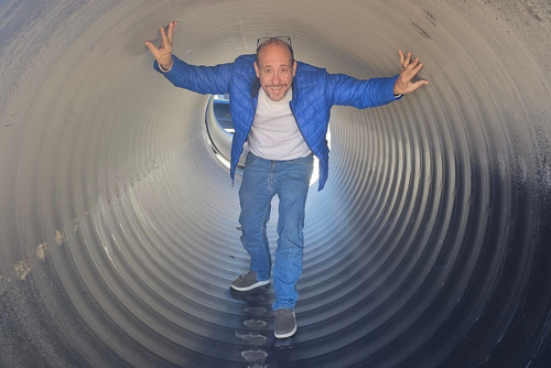 Man standing in a corrugated, cylindrical tunnel, arms outstretched. He wears a blue jacket, jeans, and sneakers.