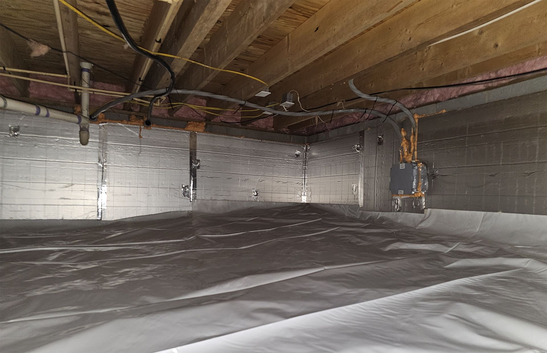 Crawlspace with a white vapor barrier, reflective insulation on walls, and exposed pipes under wooden beams.