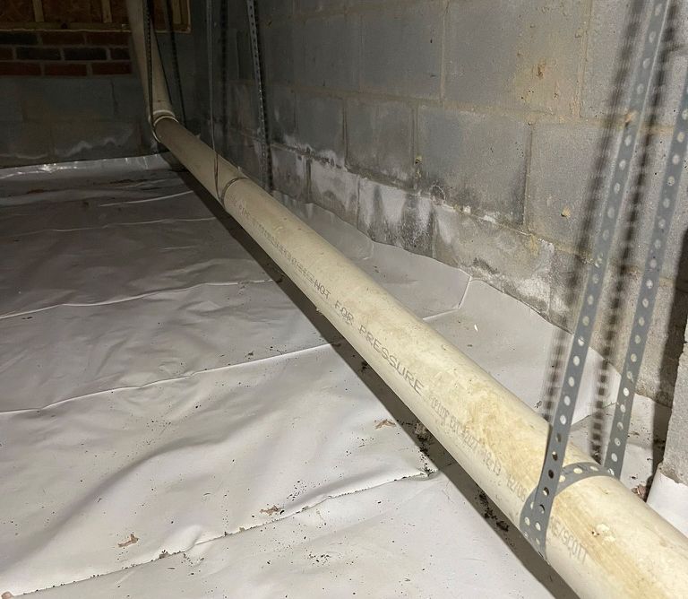 Piping supported by metal brackets, running along a concrete block wall in a basement with a white ground cover.