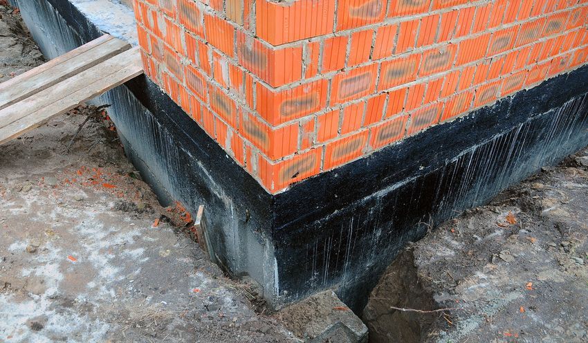 Brick wall corner on a black, waterproofed foundation in a construction setting.