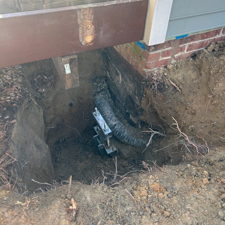 A foundation repair site with a helical pier installed beneath a house's wooden frame. 