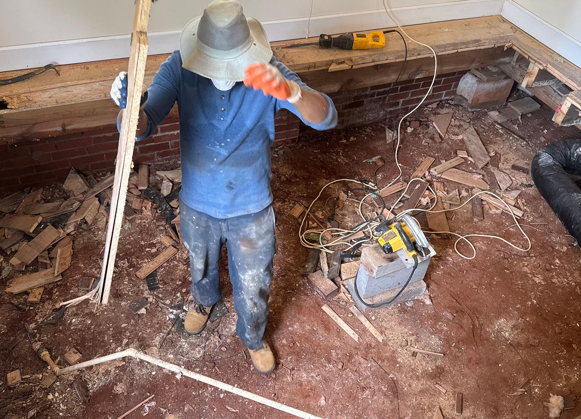 Person in a hat, working on a floor with exposed brick, holding a long strip of wood and using a saw.