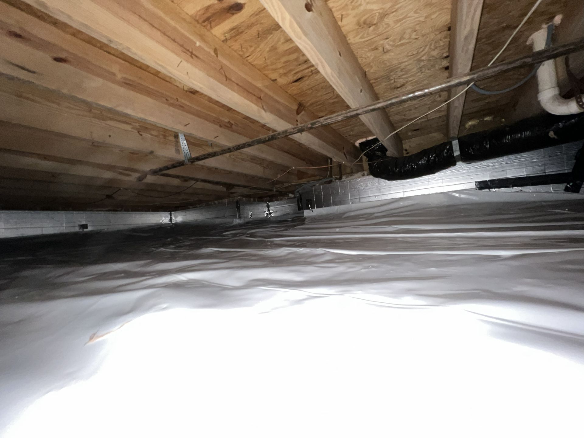 Crawl space under a wooden floor, with insulation and pipes visible.