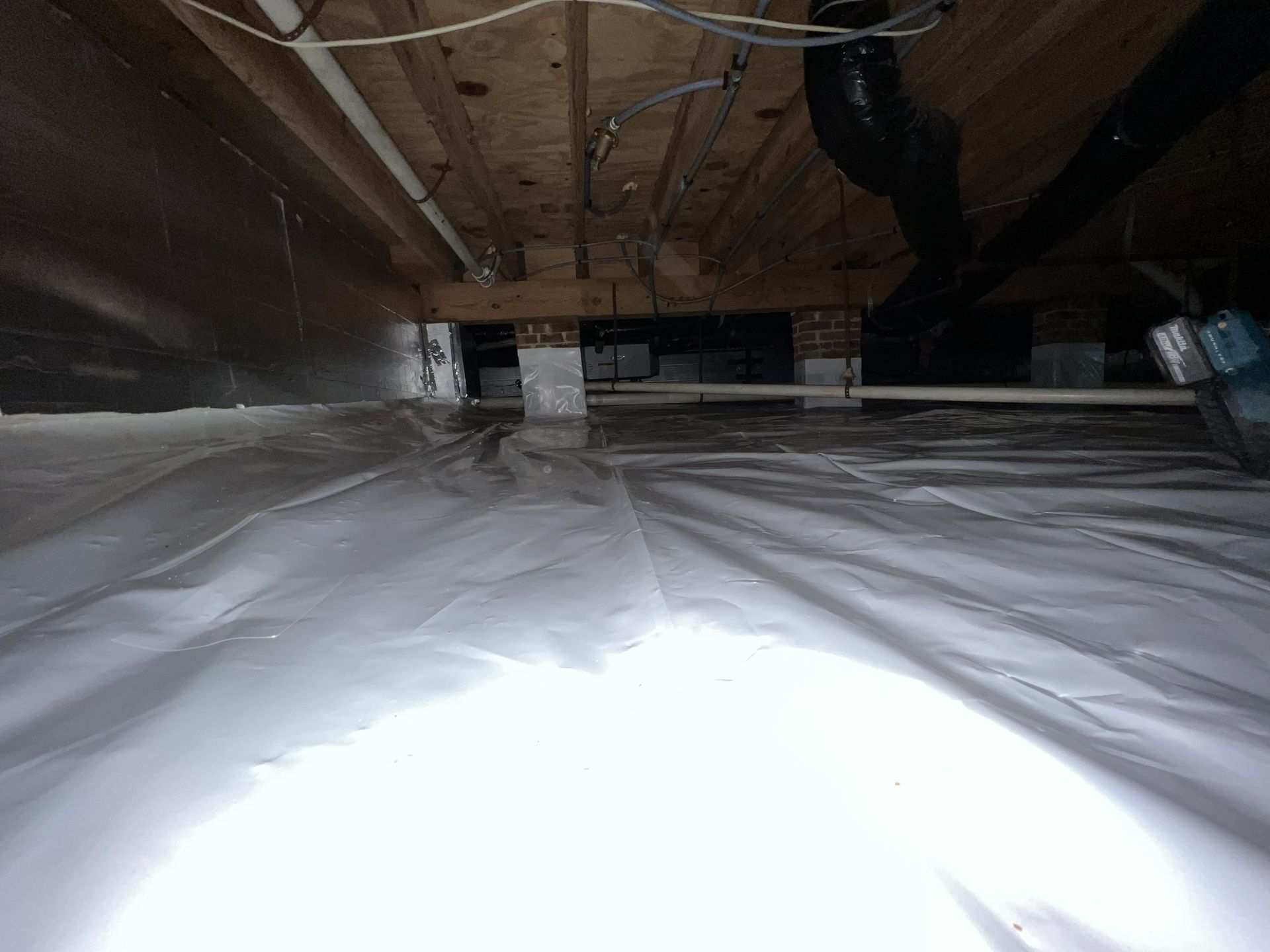 Crawl space with white vapor barrier, wood beams, and pipes.