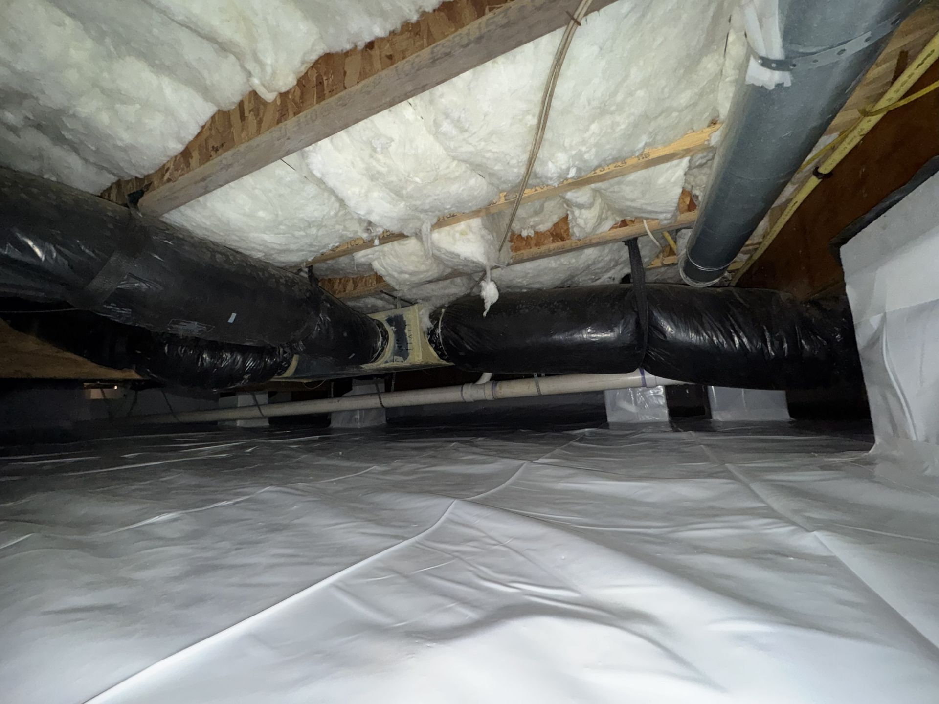 View of crawl space with white vapor barrier, insulation, and ductwork.
