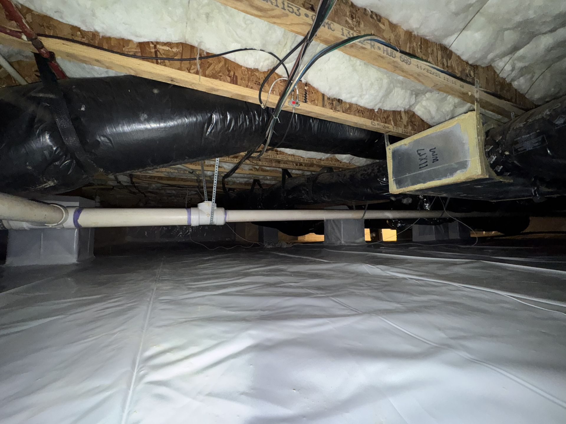 Crawlspace with white vapor barrier, gray concrete blocks supporting pipes, black insulation, and wood beams overhead.