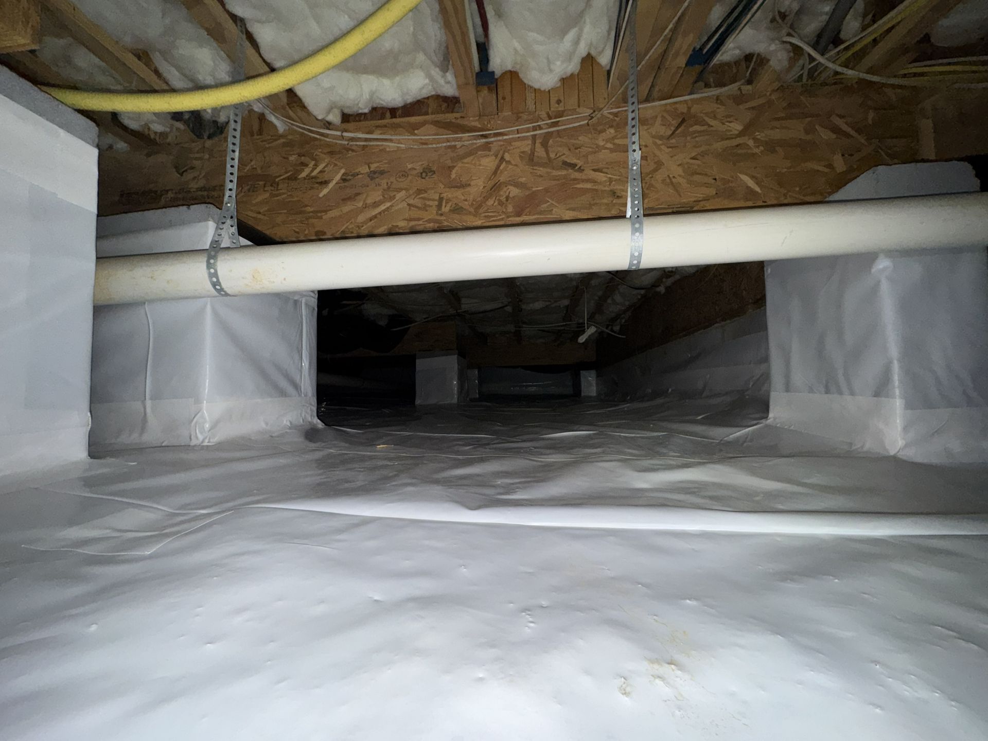 White plastic sheeting lines a crawl space, visible supports, and pipes.