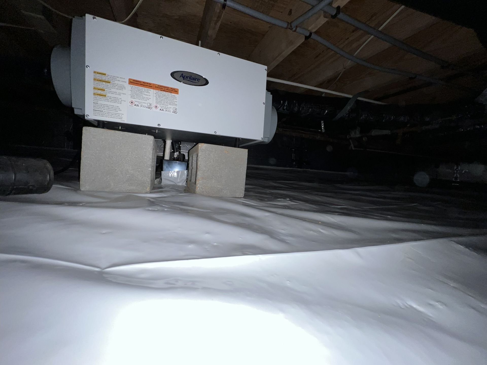 Dehumidifier in a crawl space, mounted on cinder blocks, over a white vapor barrier. Dark setting.