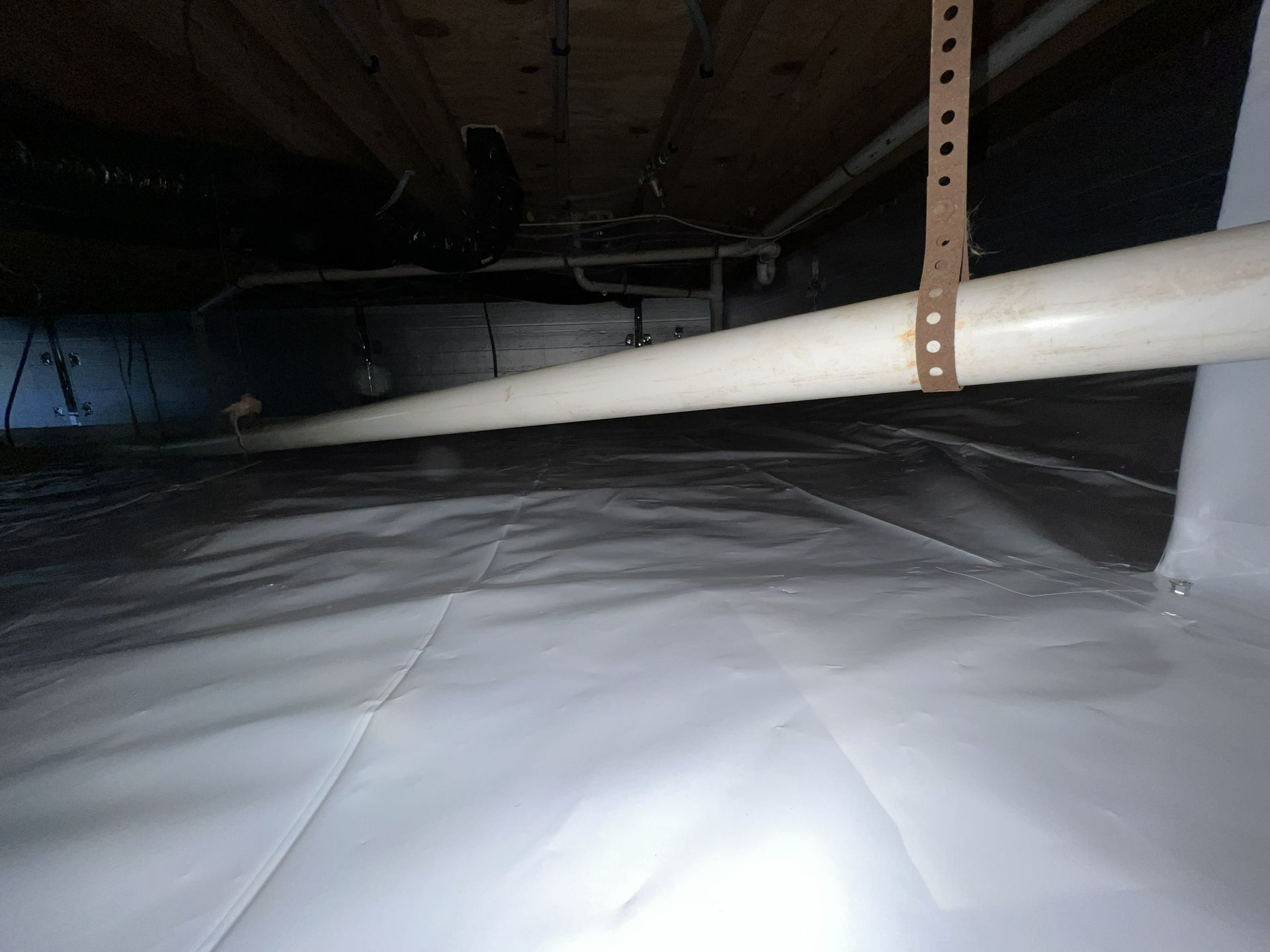 Crawl space with white vapor barrier on ground, PVC pipe, and wooden beams above.
