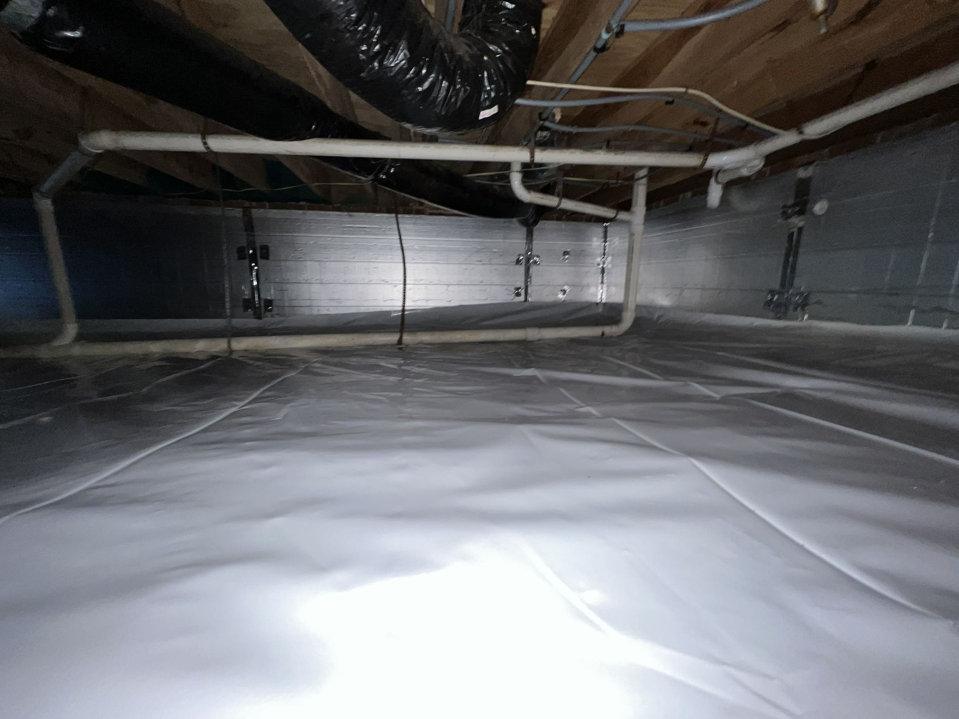 Crawl space with white plastic sheeting on the floor, pipes, and ductwork under wooden joists.