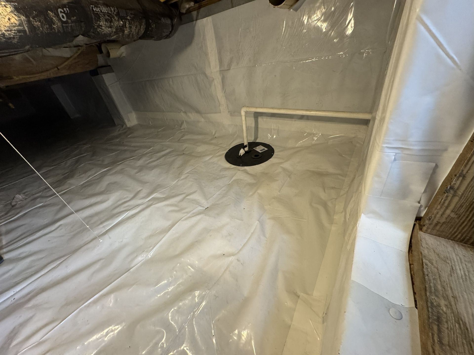 White crawl space with plastic sheeting on floor and walls, pipes, and sump pump.