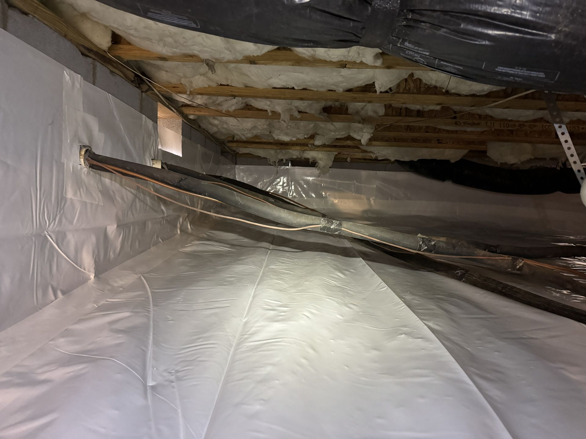 Crawlspace with white plastic sheeting, exposed wood beams, and insulation.  A dark tube runs across the space.