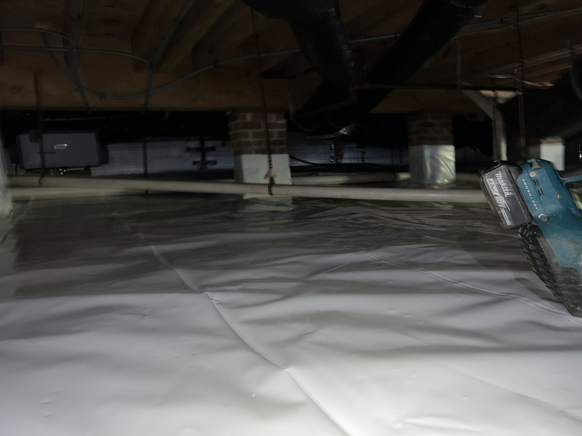 White crawlspace with plastic ground cover, support beams, and plumbing.