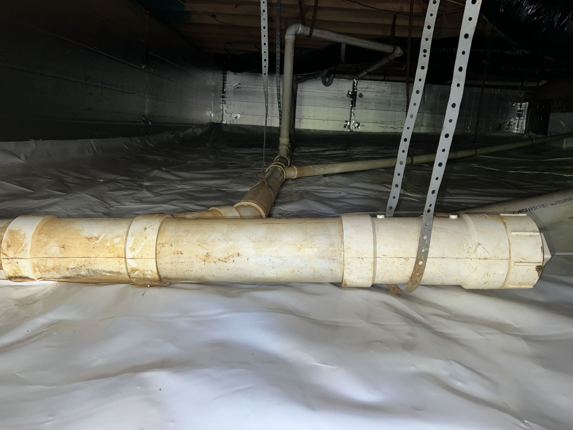 White PVC pipes with rust stains, set in a crawlspace with white vapor barrier, metal straps, and ductwork.