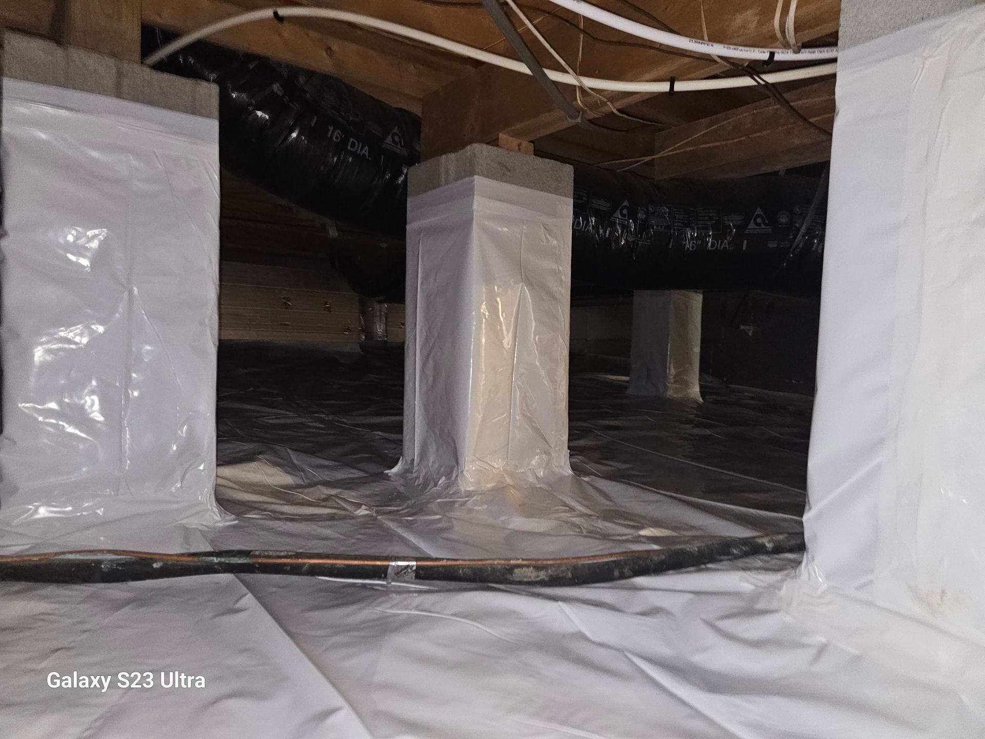 Crawl space with white plastic sheeting on the floor and supporting pillars; black insulation tubes overhead.