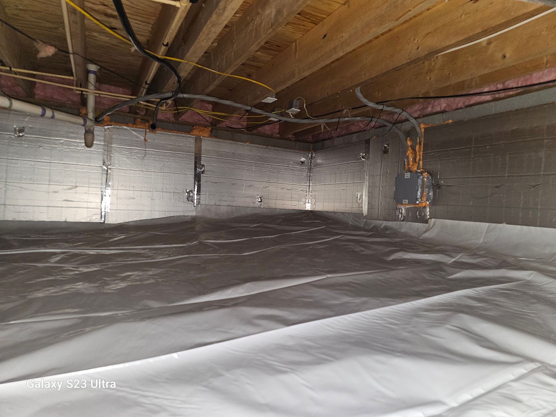 Crawl space with white ground cover and silver insulated walls under a wooden ceiling with pipes and wires.