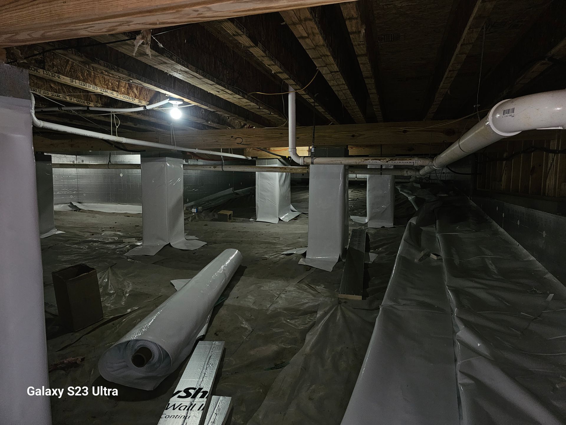 Crawlspace with support posts, insulation, and pipes. The space is dimly lit and appears unfinished.