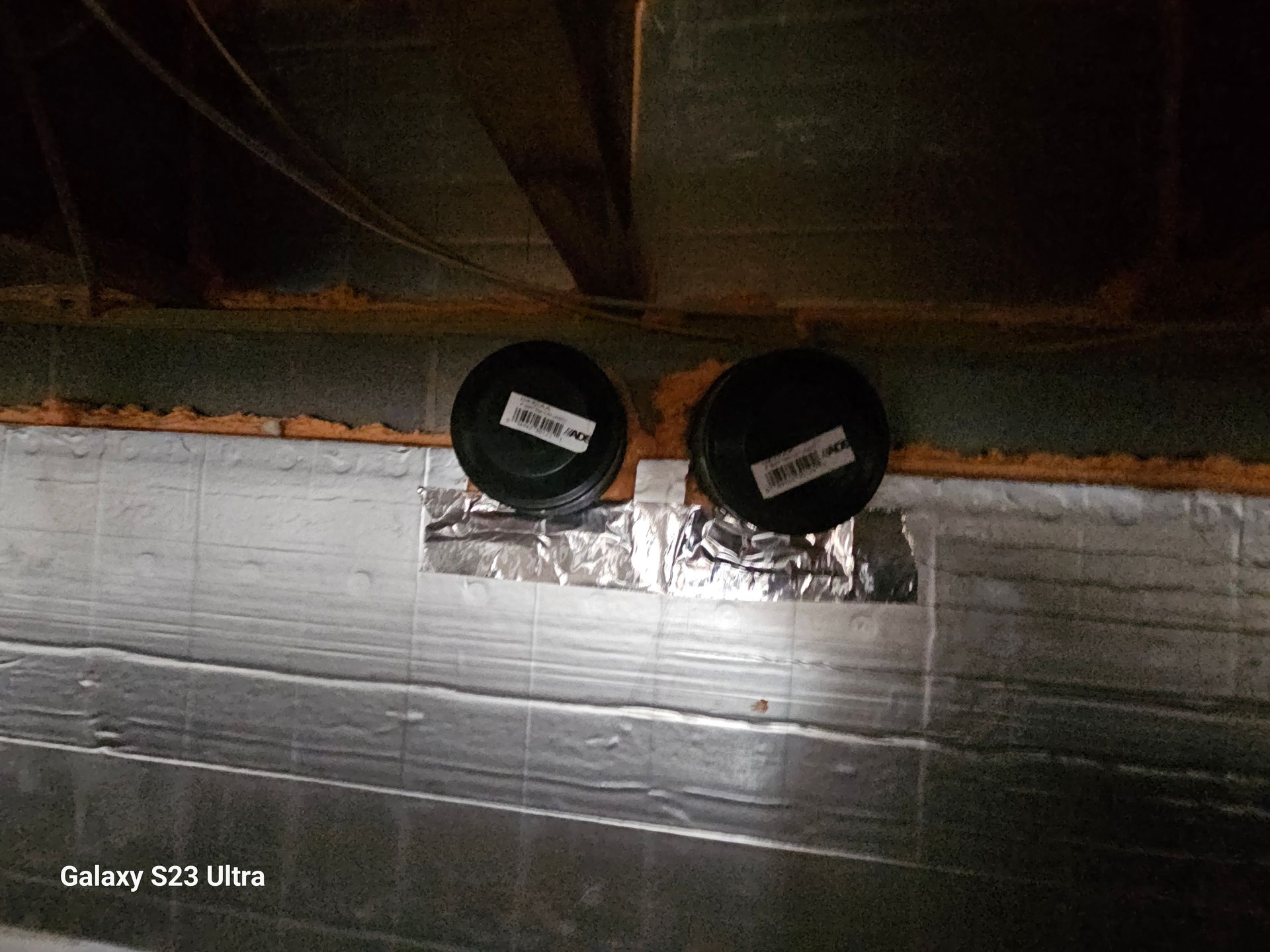 Two black round vents on foil tape in a dark basement. Silver insulation covers the wall.