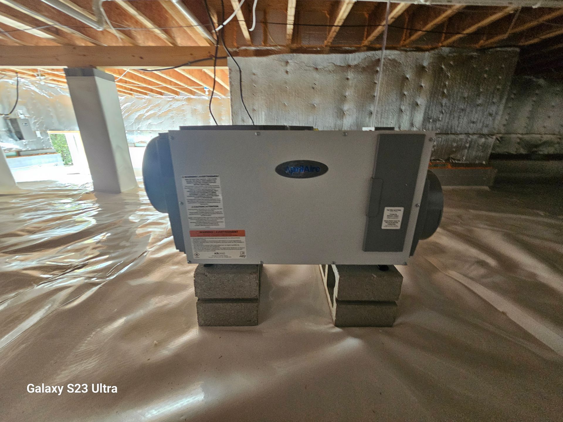 A gray dehumidifier sits on concrete blocks in a crawl space with white plastic sheeting.