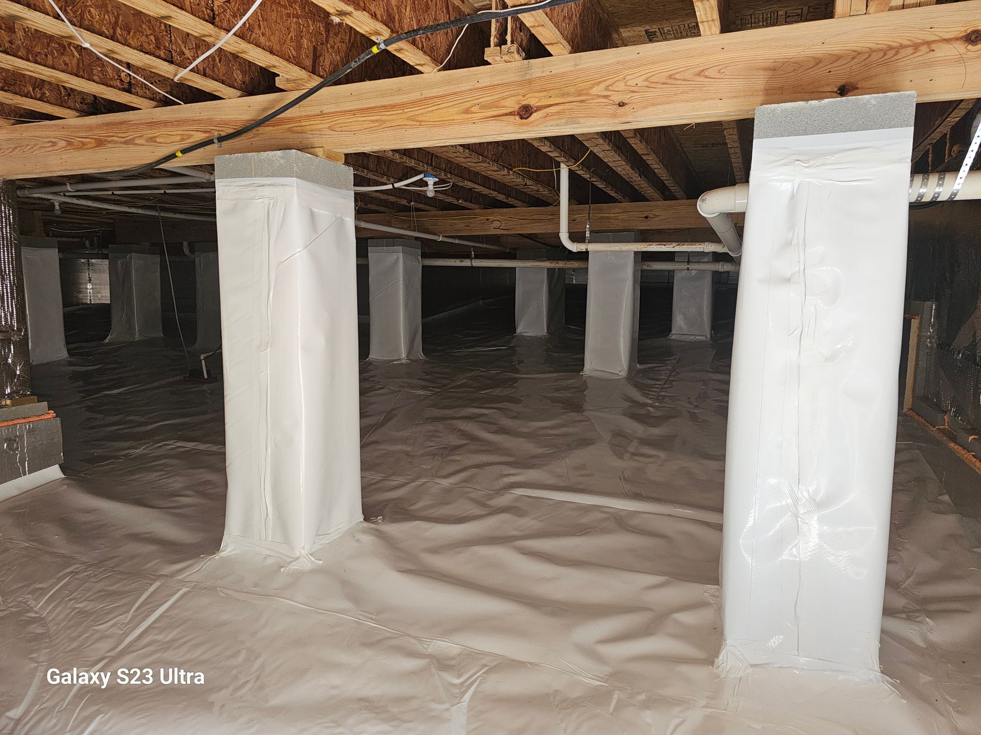 Crawlspace with support posts wrapped in white, covered in a white vapor barrier. Wooden beams above.