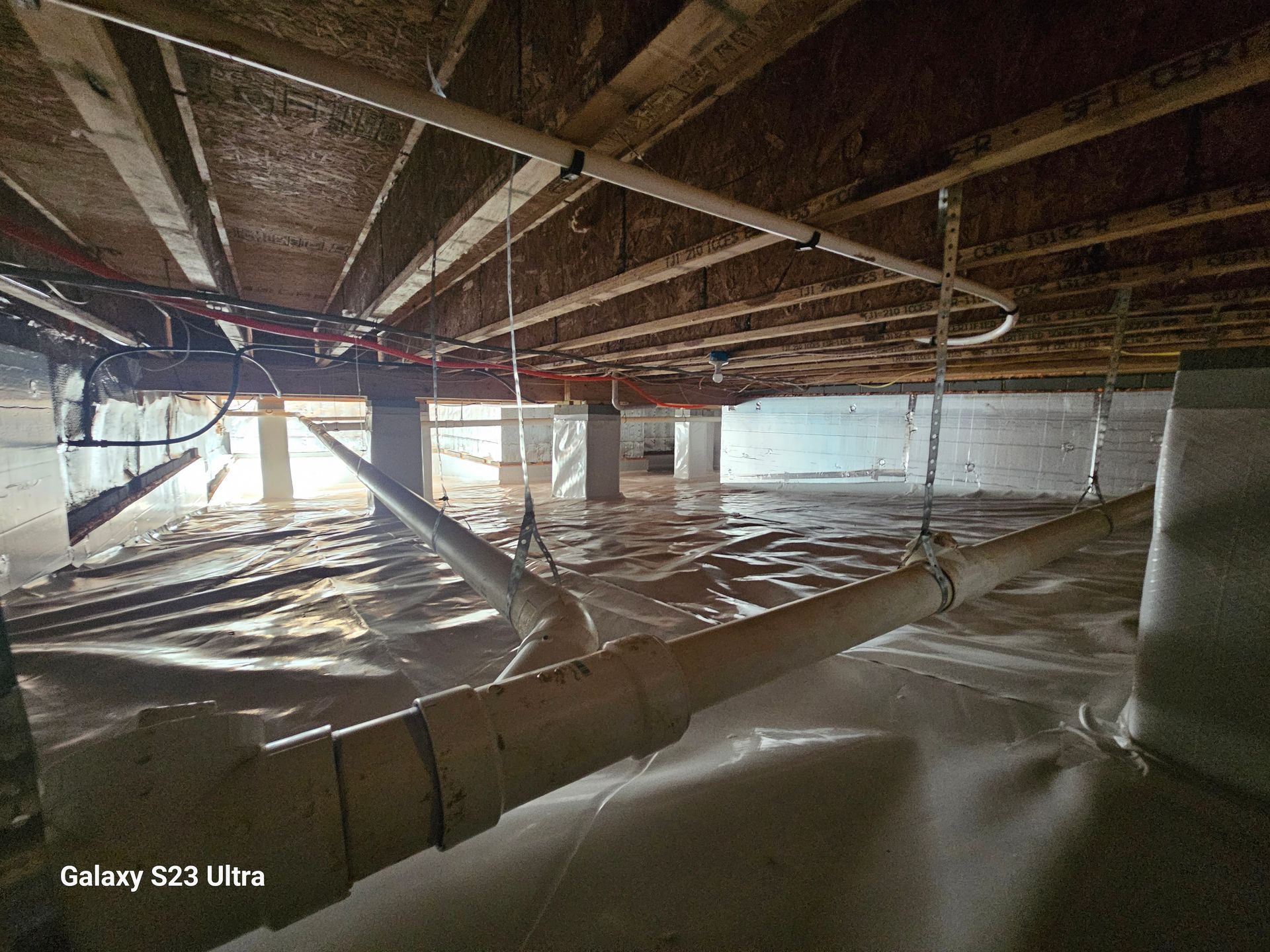 Underneath a building, a flooded crawlspace with pipes, supports, and plastic sheeting visible.