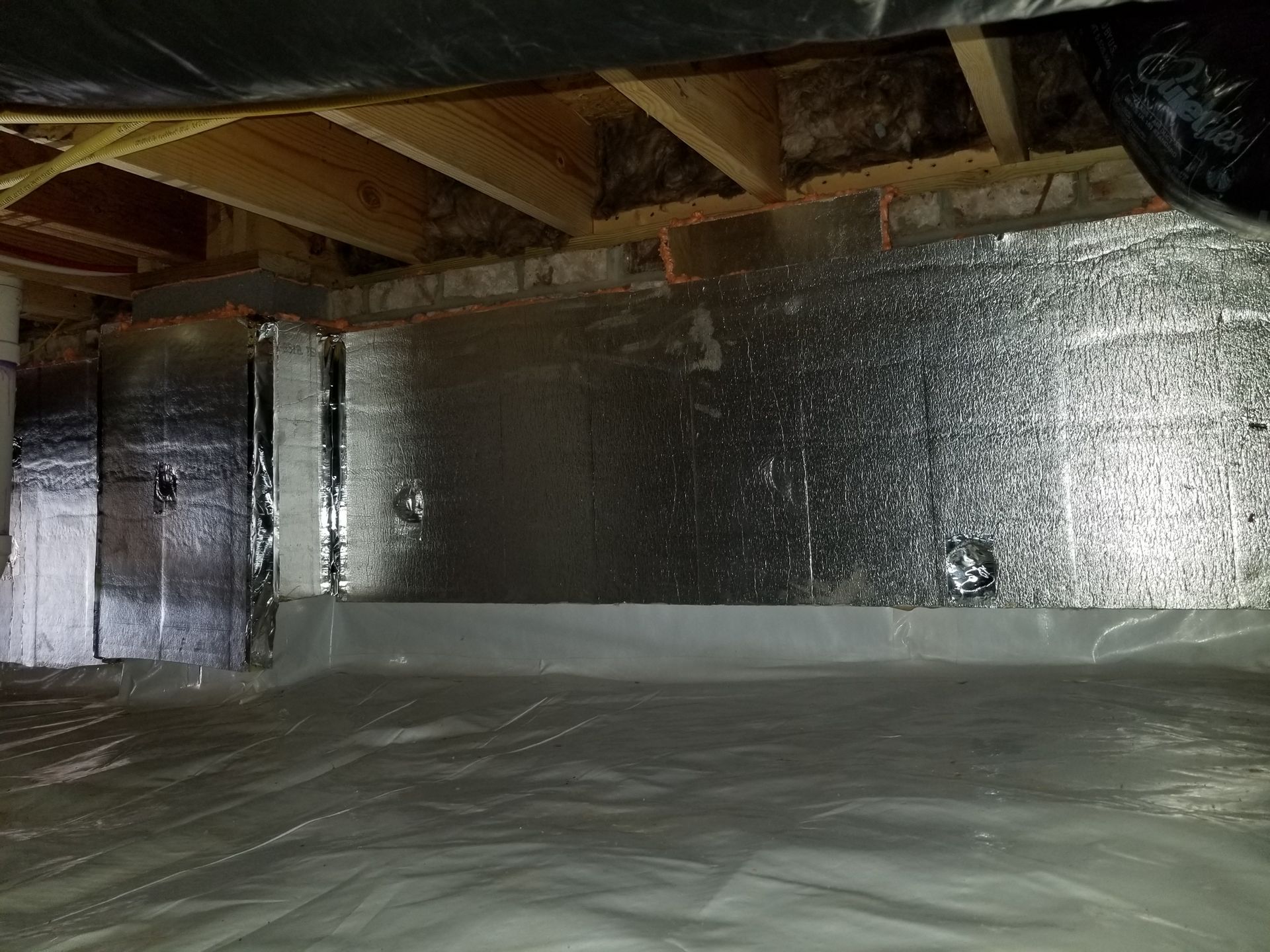 Crawl space with silver insulation on walls and a vapor barrier on the ground. Wooden beams are visible above.