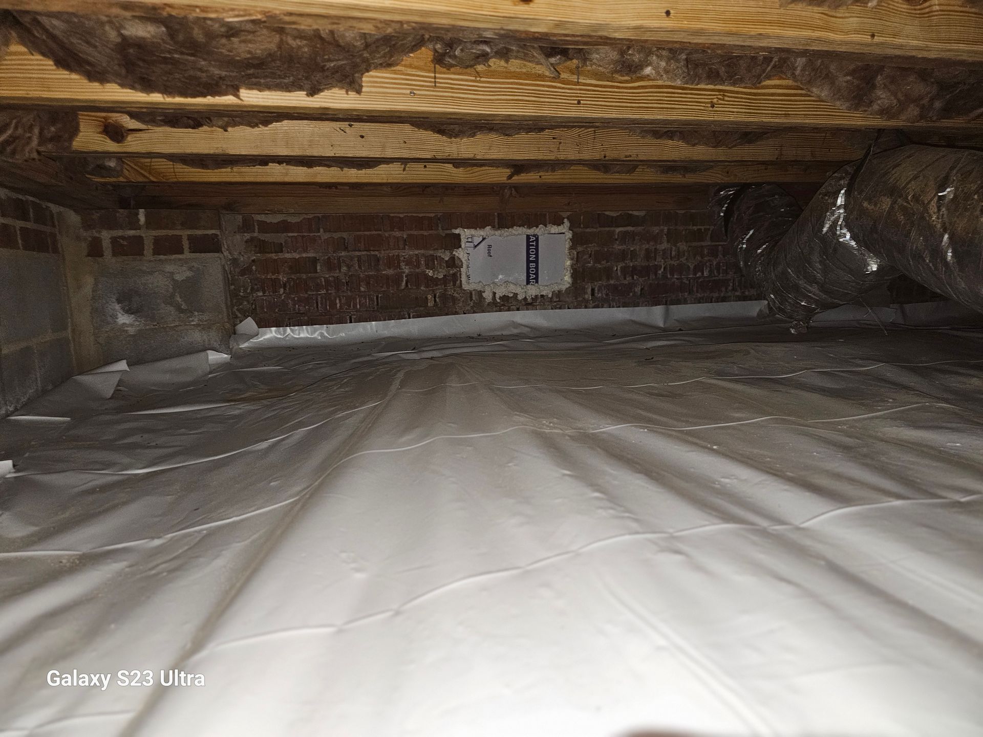 Crawlspace with white vapor barrier covering the ground. Brick foundation, wooden beams, and ductwork are visible.