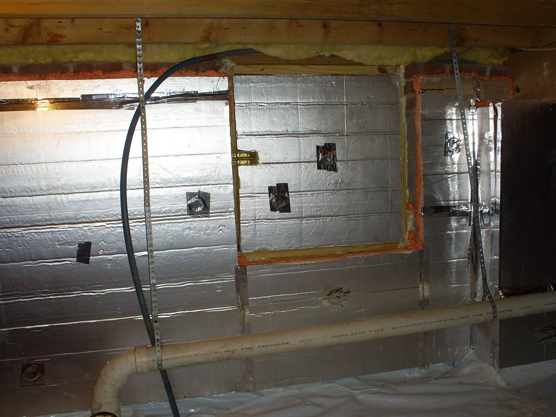 Interior view of a crawl space with insulated walls, electrical outlets, and pipes. Silver insulation and wood framing are visible.