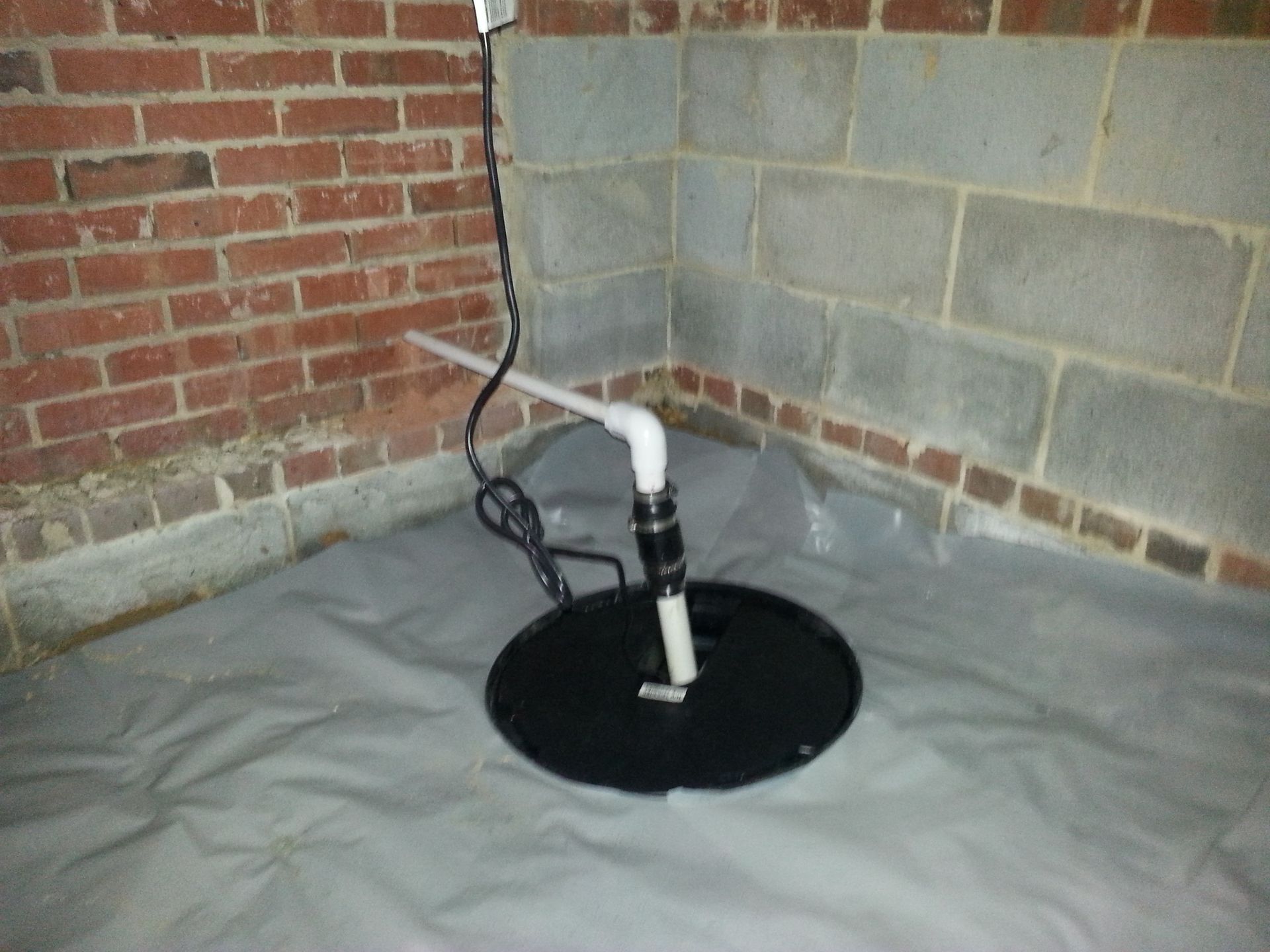 A basement sump pump with PVC piping.  The pump is black, set on gray plastic, near brick and concrete block walls.