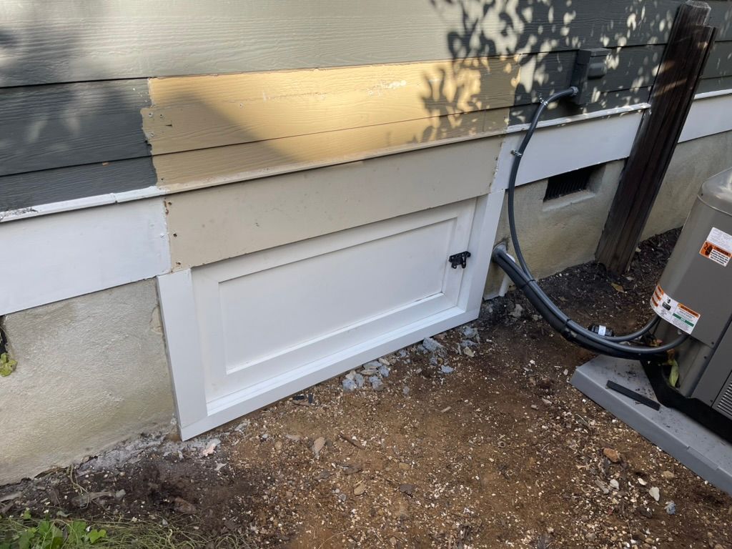 White cellar door on a tan house exterior, beside a black hose and air conditioning unit.