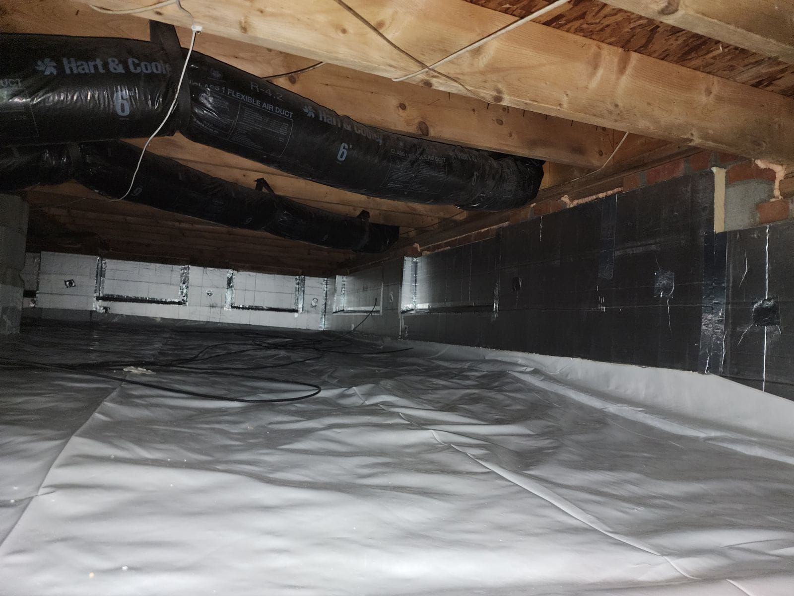 Crawlspace with insulation and vapor barrier, exposed wooden beams and ductwork overhead.