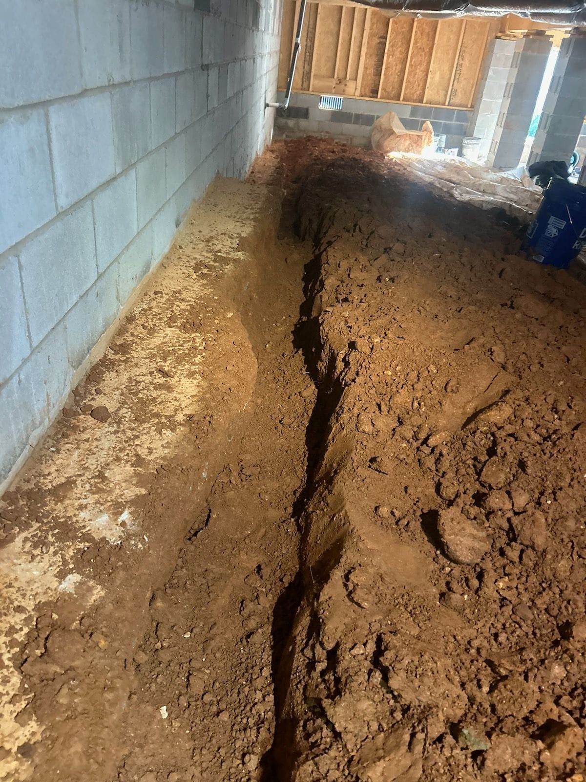 Trench dug along a concrete block wall inside a building. Dirt is reddish-brown.
