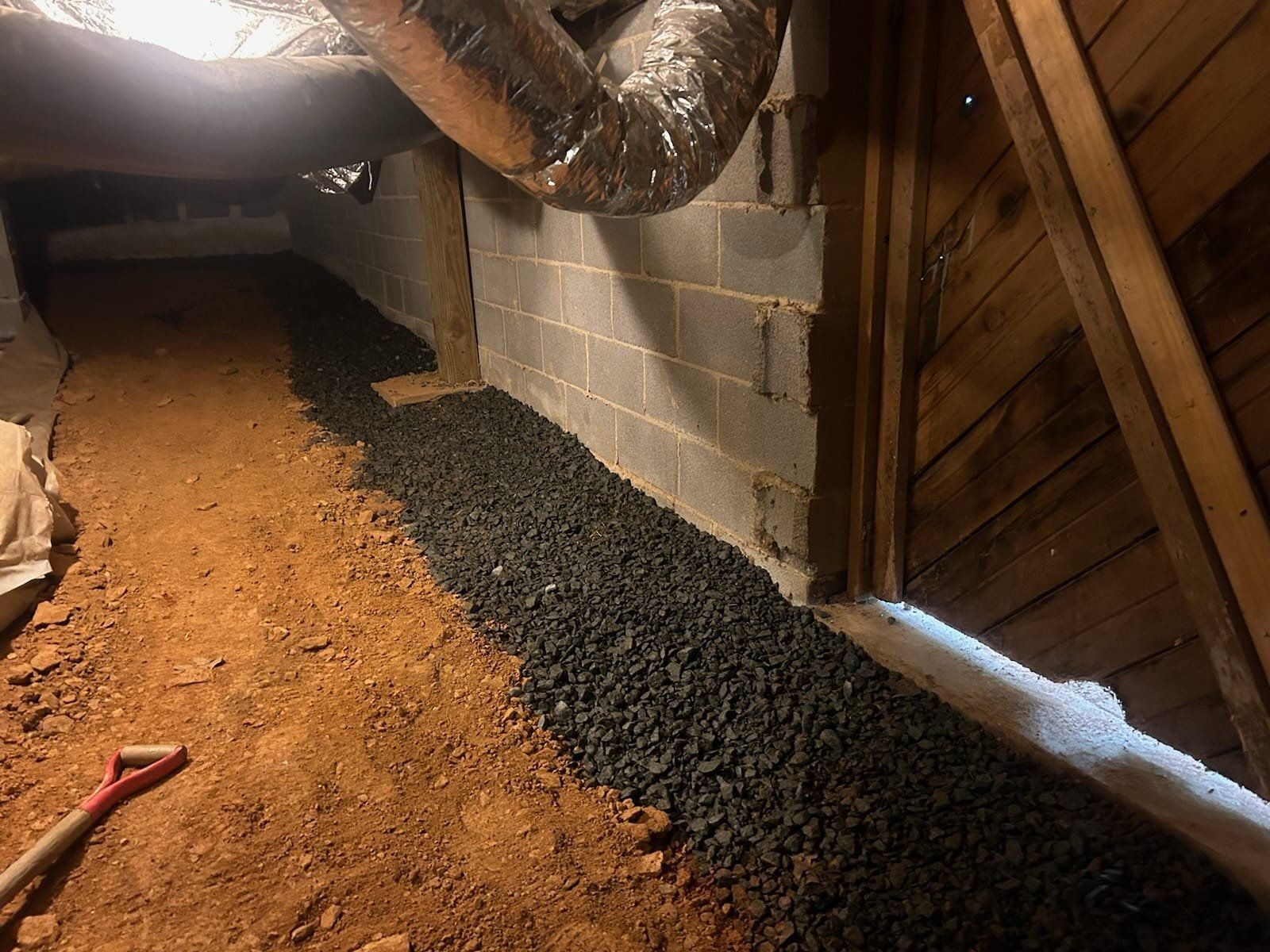 Crawlspace with dirt floor and black gravel along a cinder block wall, a duct, and wooden doorframe. A shovel lies on the dirt.