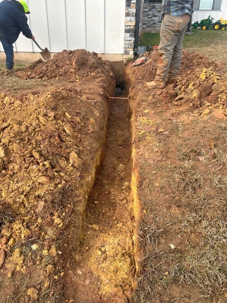 Two people digging a narrow trench in brown earth next to a white building.