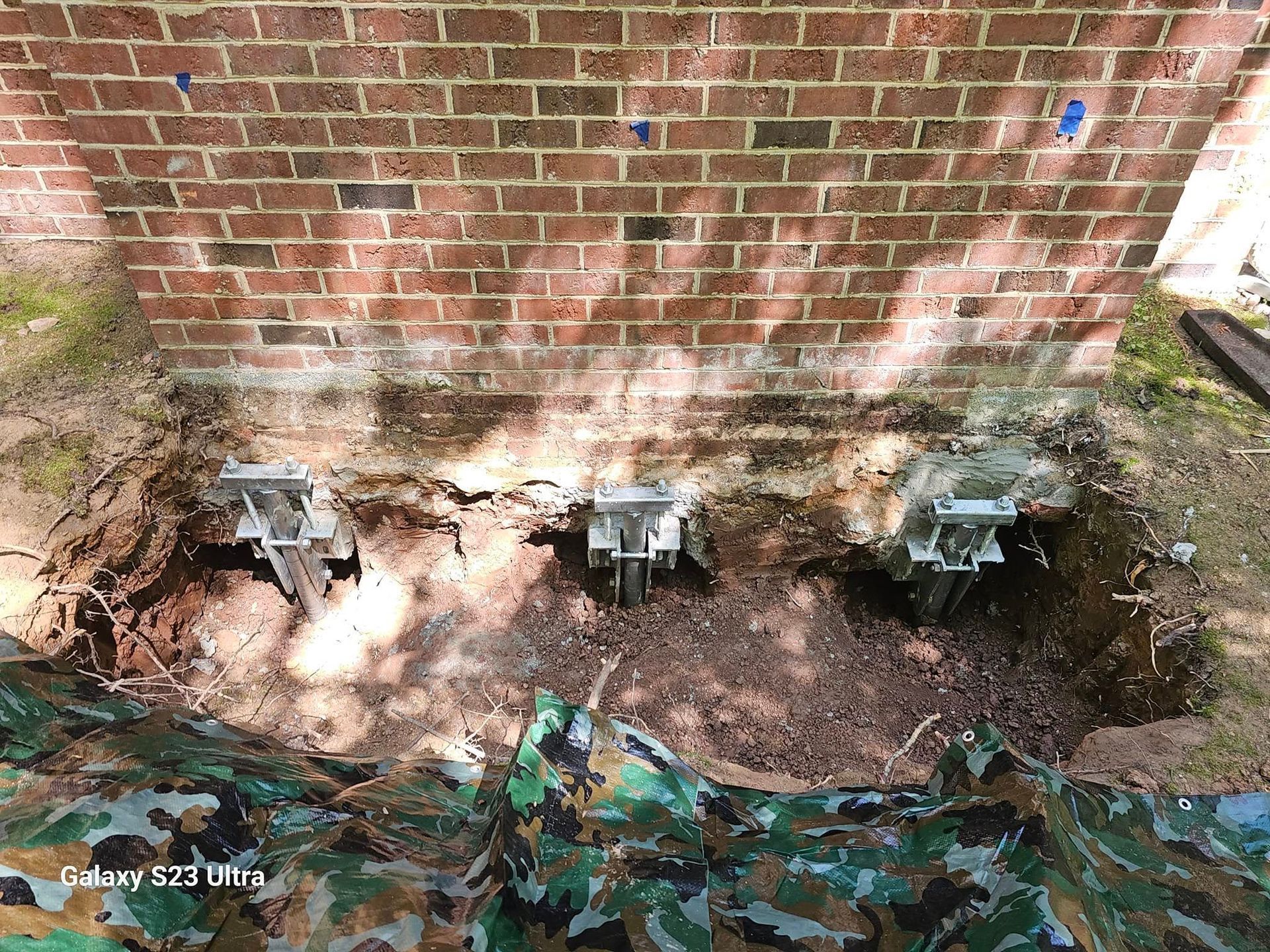 Brick wall with three metal foundation piers installed in dug-out dirt, camouflage fabric in front.