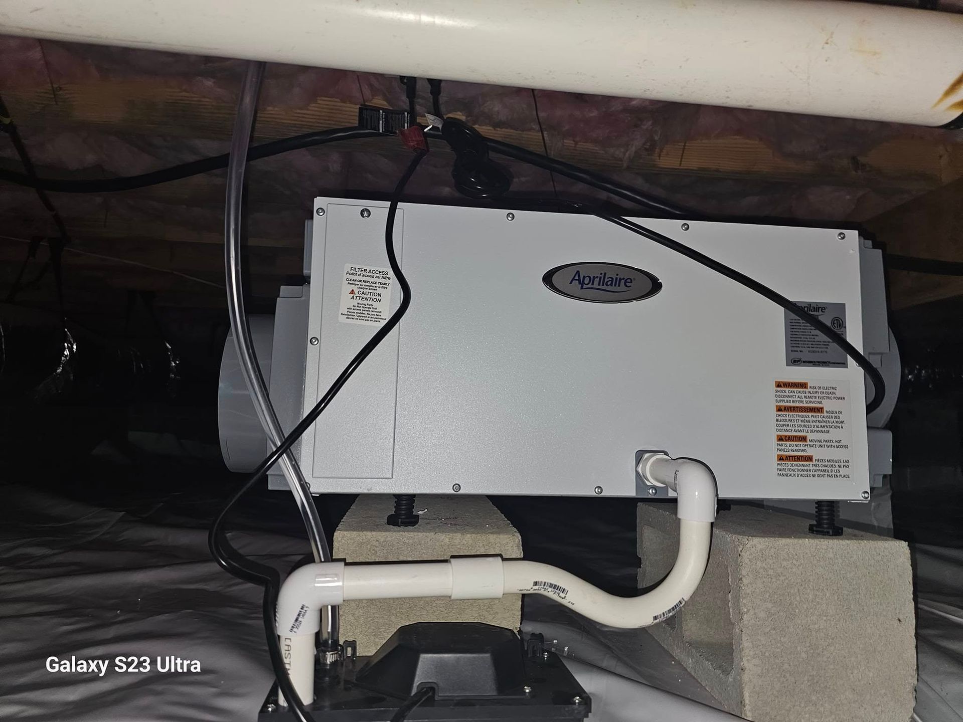 Dehumidifier in a crawlspace, mounted on cinder blocks, with drain tubing and electrical wiring.