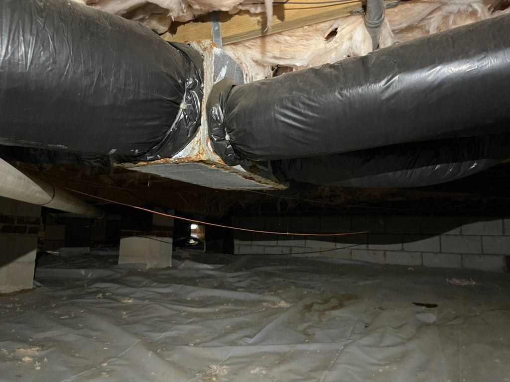 View of a crawl space with black HVAC ducting, insulation, and moisture barrier on the ground.
