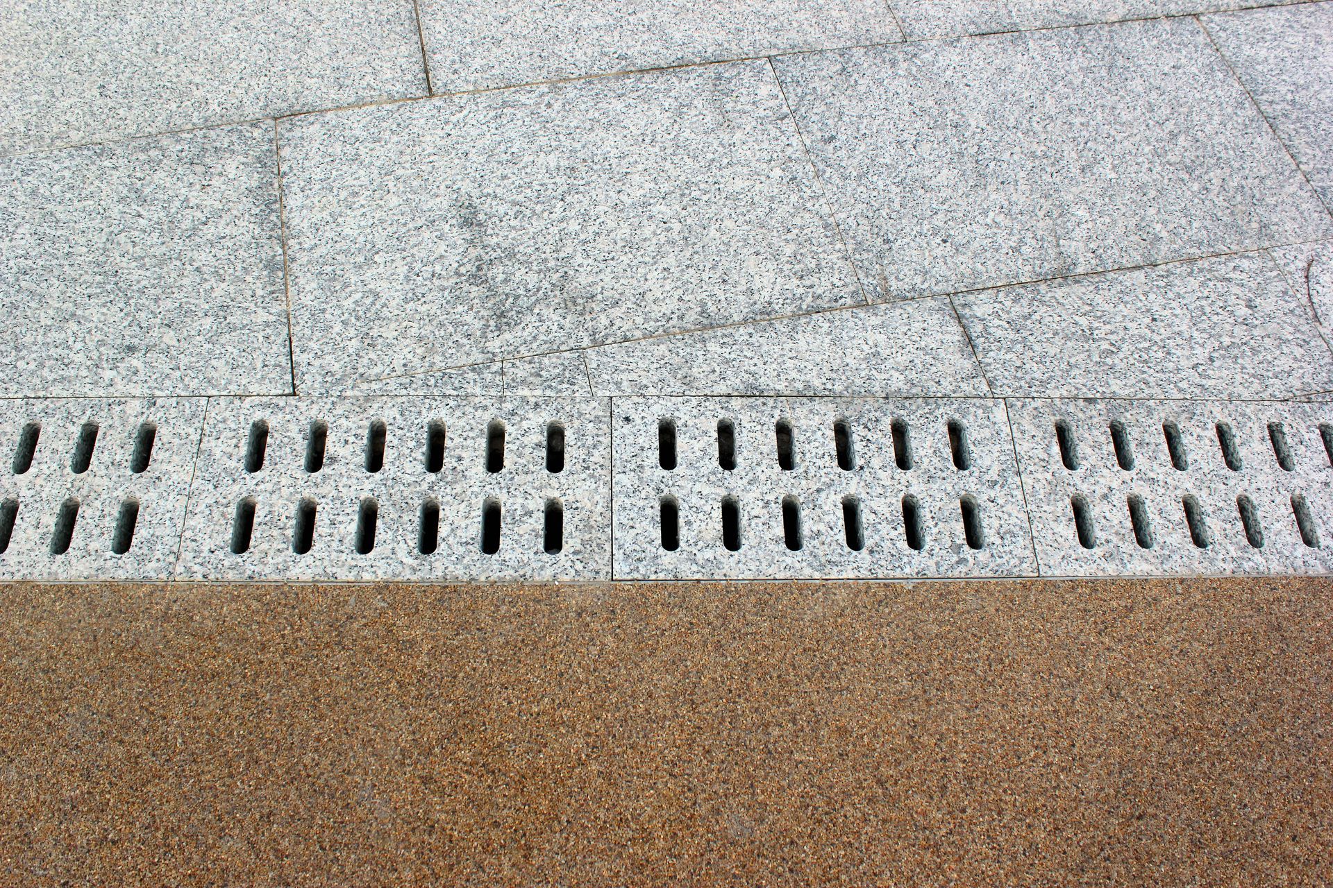Granite paving and drain cover meet with a brown textured surface.