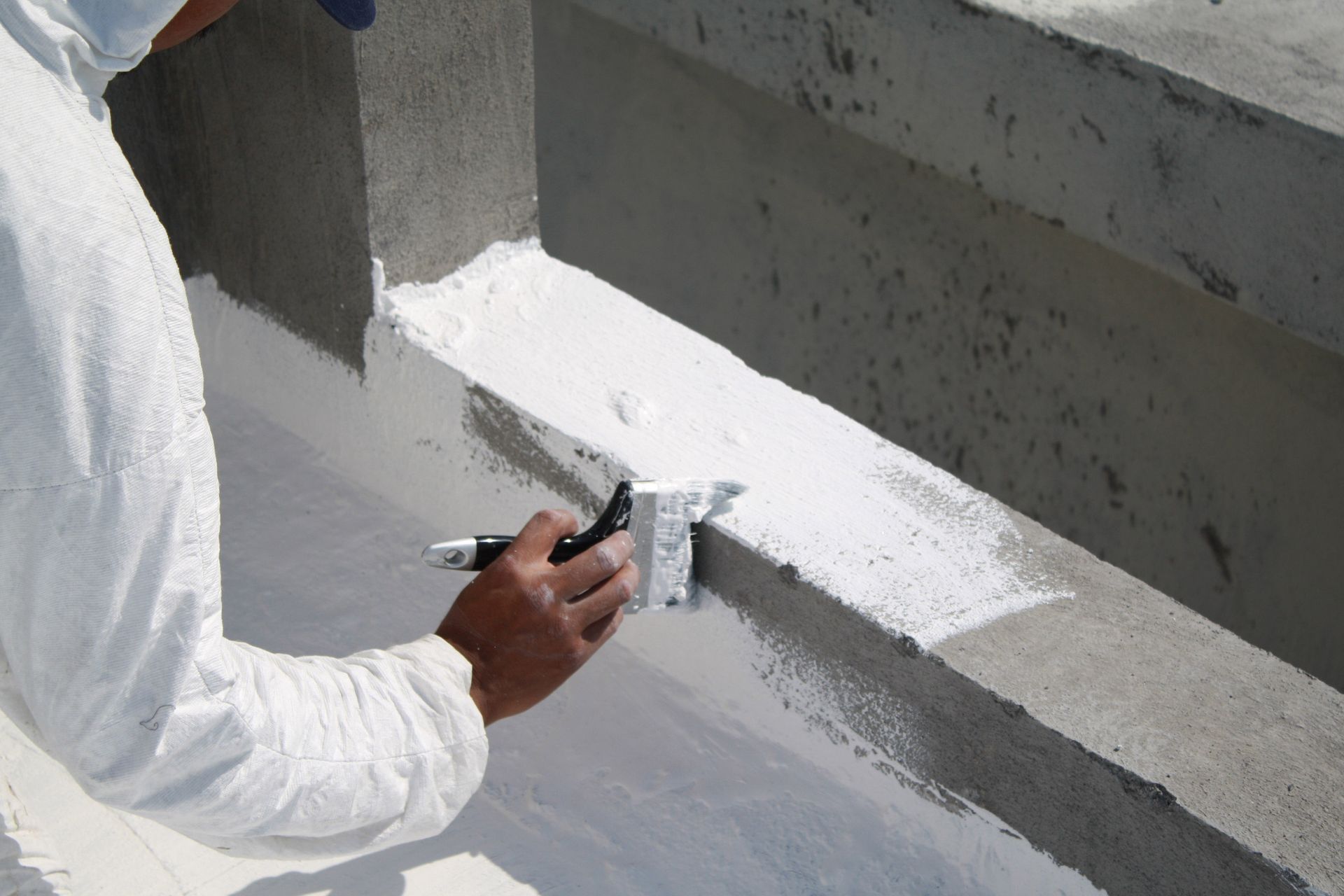 Person in white protective suit painting a white sealant on a concrete structure.
