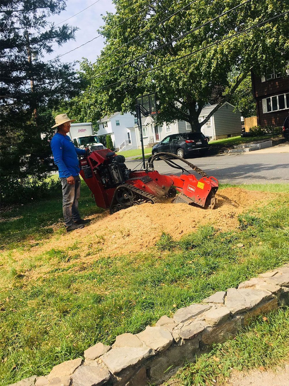 Worker operating a red excavator on a grassy street-side mound near trees and houses