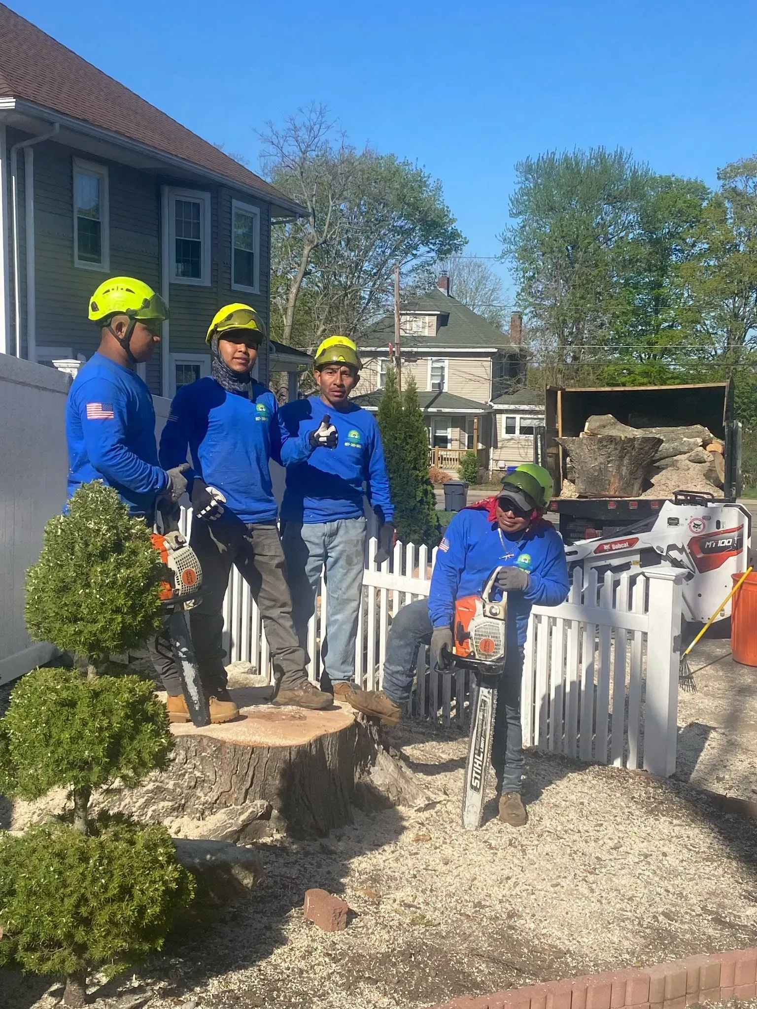 Five workers in blue jackets and yellow hard hats by a white fence in a garden with a house and gravel path.