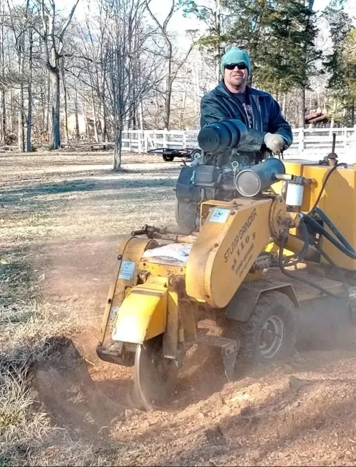 Man in blue jacket riding a yellow construction vehicle through a dirt path, kicking up dust