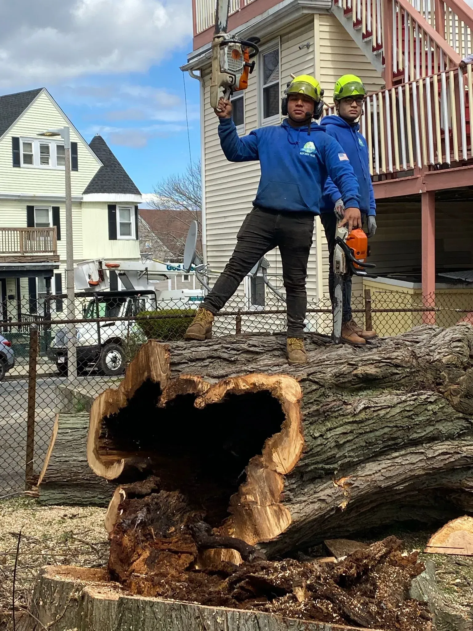 Two workers in safety gear stand beside a huge fallen tree trunk on a residential street.